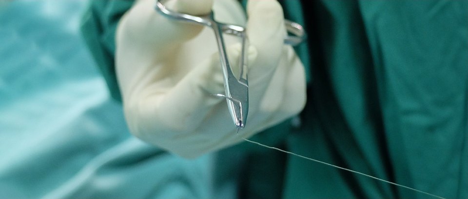 짙은 청록 수술포를 배경으로 장갑 낀 손이 지혈겸자로 봉합사를 팽팽히 당기는 수술 장면 클로즈업