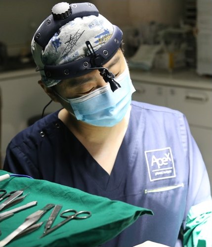 수술등 헤드램프와 마스크를 착용한 남성 집도의가 수술복 차림으로 초록색 소독포 위 수술기구를 내려다보는 모습