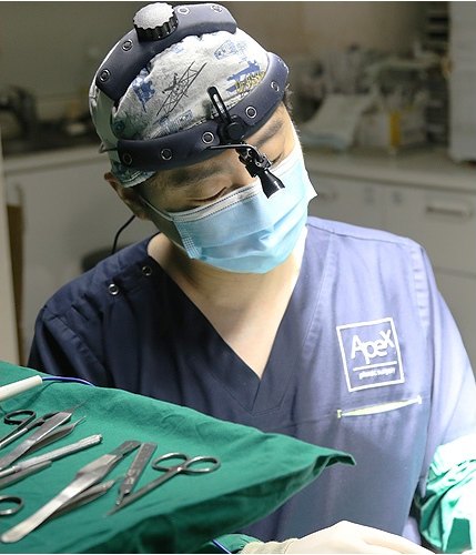 수술모와 확대경, 마스크를 착용하고 수술에 임하는 에이펙스성형외과 집도의. 앞쪽 초록 소독포 위에 수술 기구가 놓여 있다