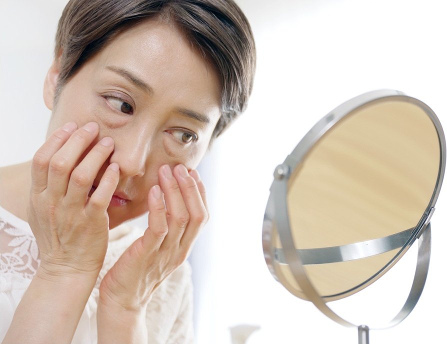 중년 여성이 두 손으로 눈 밑을 만지며 탁상 거울을 들여다보는 사진