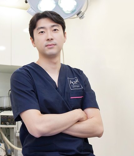 네이비 수술복을 입고 팔짱을 낀 남성 의료진이 수술실 무영등 아래에서 정면을 바라보는 인물 사진