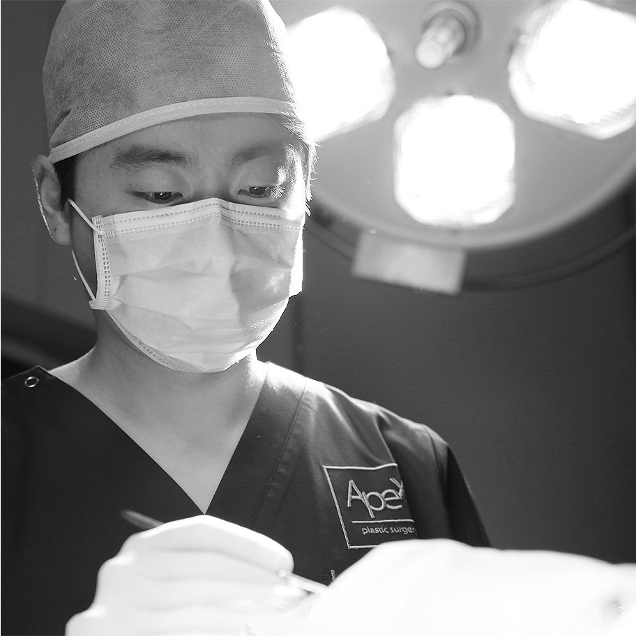 어두운 수술복을 입은 에이펙스성형외과 의료진이 수술 기구를 쥐고 있는 흑백 클로즈업. 가슴에 Apex plastic surgery 자수 로고가 보인다.