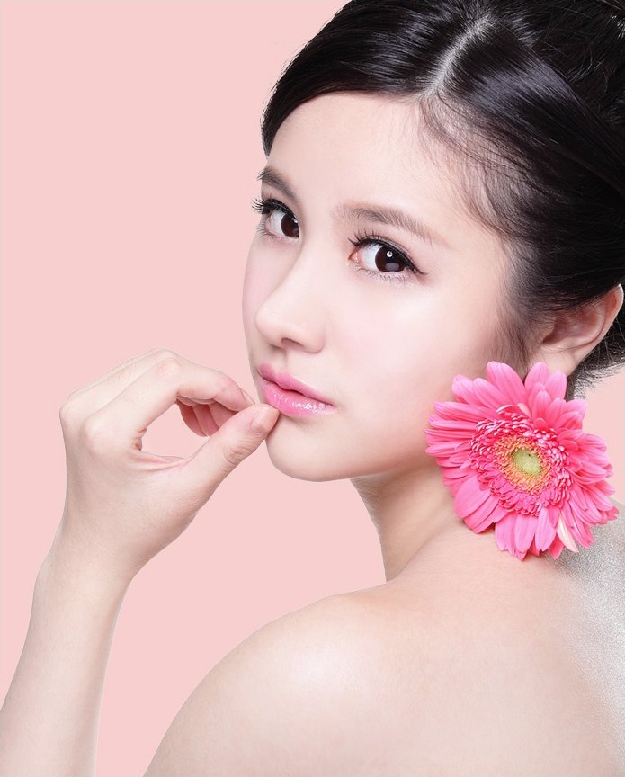 연분홍 배경 앞에서 거베라 꽃을 어깨에 두고 턱에 손을 얹은 여성의 피부 클로즈업