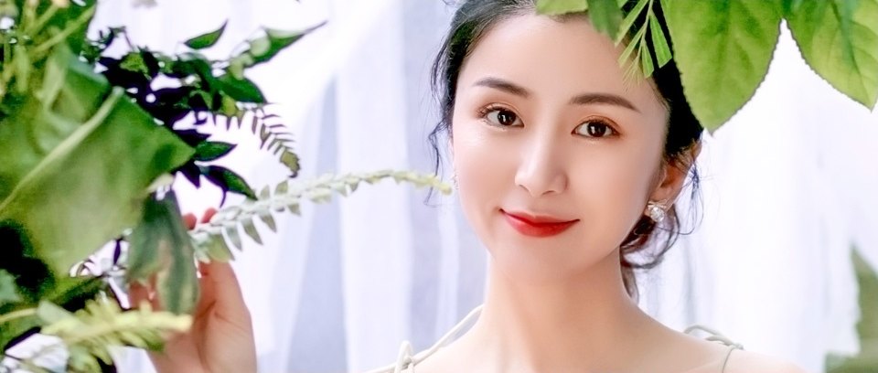 초록 잎사귀 사이에서 가지를 손으로 잡고 정면을 바라보며 옅게 미소 짓는 젊은 여성의 상반신 클로즈업