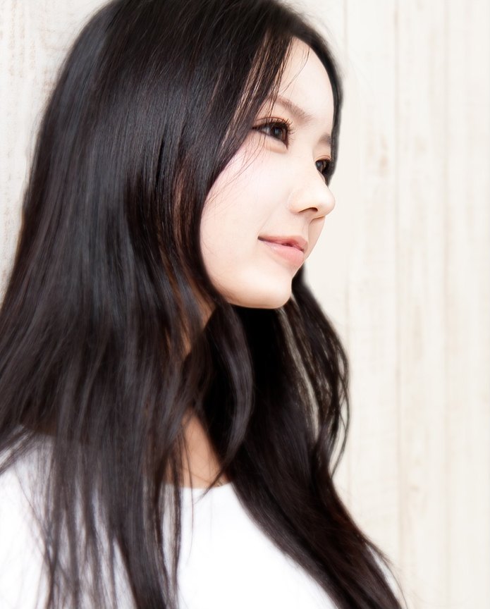 긴 흑발의 젊은 동양인 여성이 밝은 우드 패널 벽 앞에서 오른쪽을 바라보는 반측면 상반신 컷