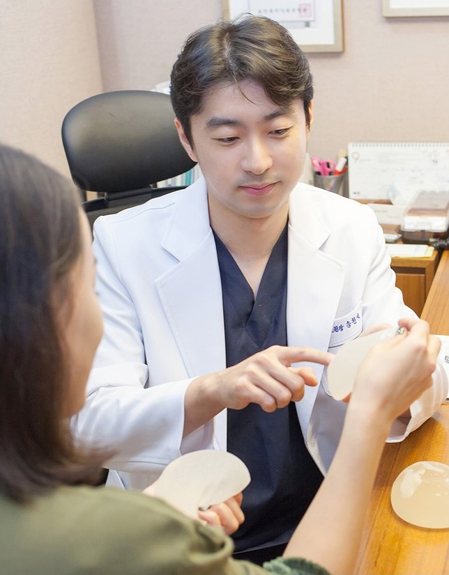 흰 가운을 입은 의사가 진료실에서 원형 실리콘 유방 보형물을 들어 보이며 내원객과 상담하는 장면