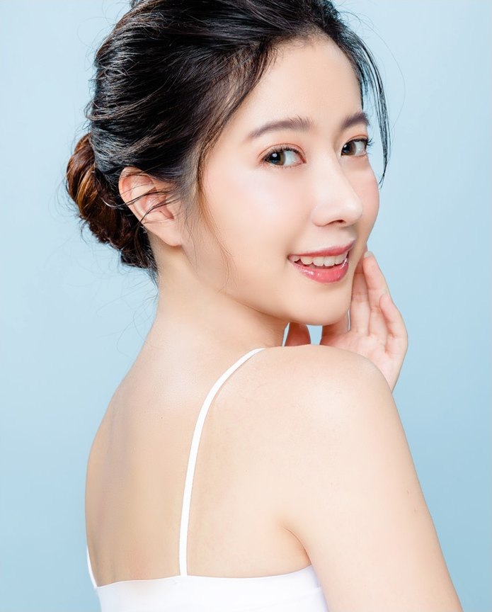 머리를 올려 묶은 여성 모델이 어깨 너머로 돌아보며 미소 지으며 손끝을 뺨에 가볍게 대고 있는 모습